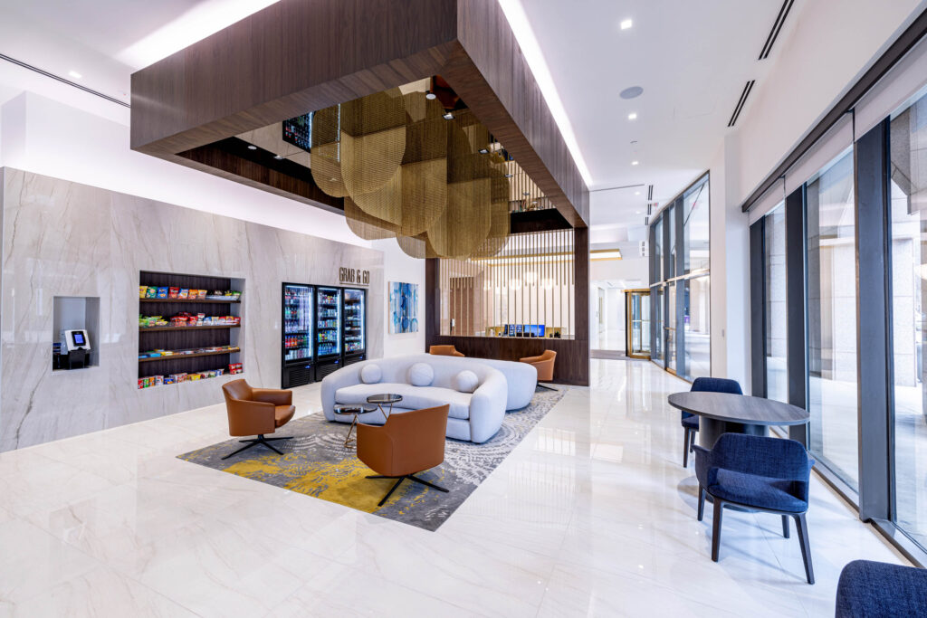 Modern lounge in a plaza setting, with a circular sofa, brown chairs, and snacks against large windows and bright lighting.