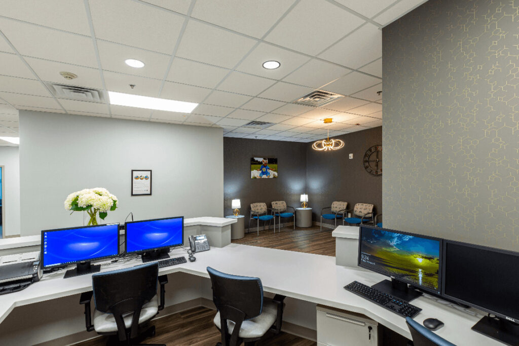 Modern office reception with curved desk, two monitors showcasing technology, chairs, decorative wall, and ceiling lights.