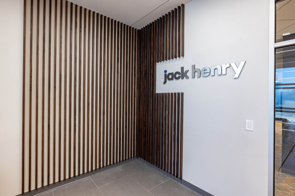 Wood-paneled wall corner with a jack henry sign in metallic letters on the right, adjacent to a modern office area.