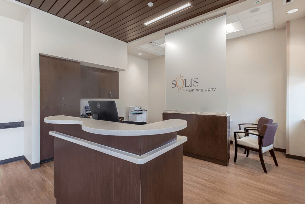 Empty clinic reception area with a wooden desk, chairs, and a sign saying Solis Mammography Radiology on the wall.