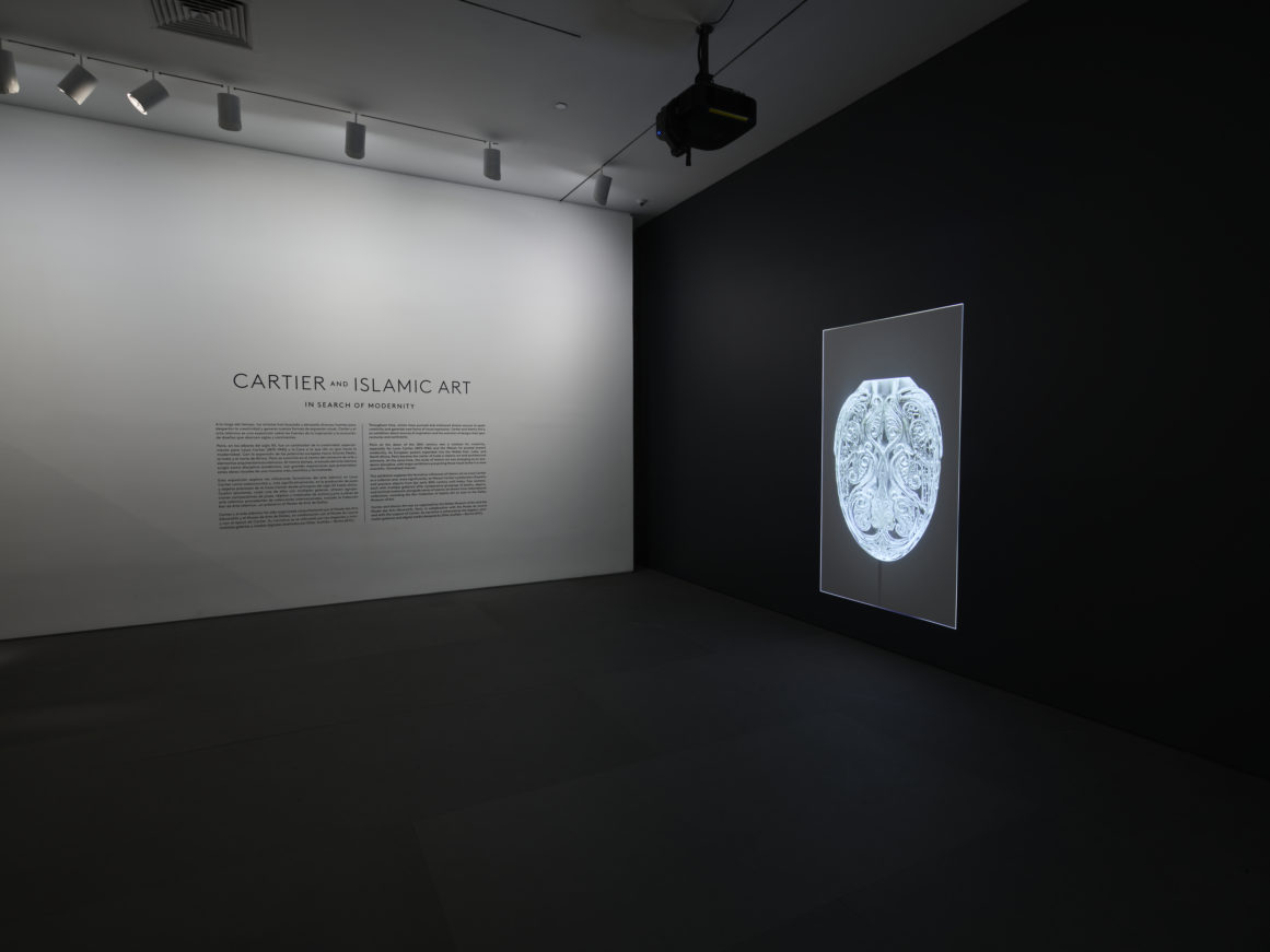 Dimly lit exhibition room displaying a wall text about Cartier and Islamic art, with a glowing framed artwork on the right wall.