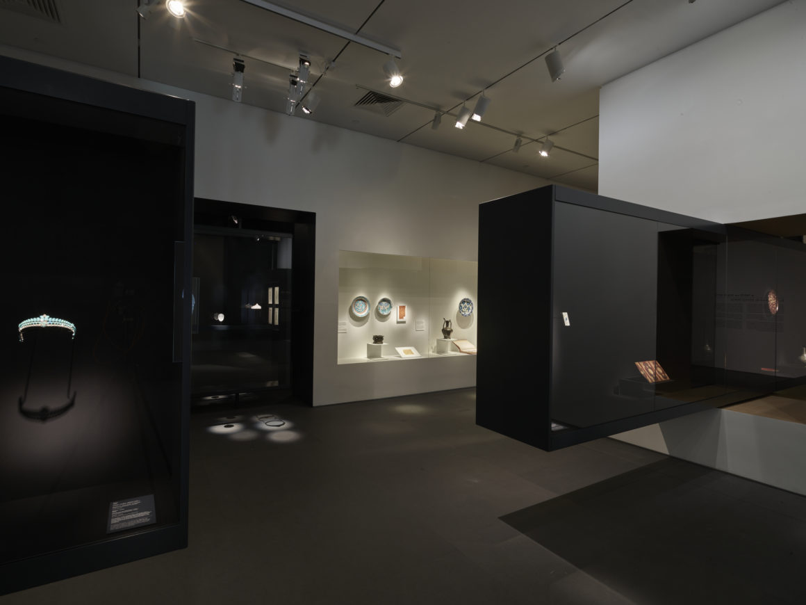 Dimly lit museum exhibit with various artifacts displayed in glass cases, including jewelry, pottery, and books.