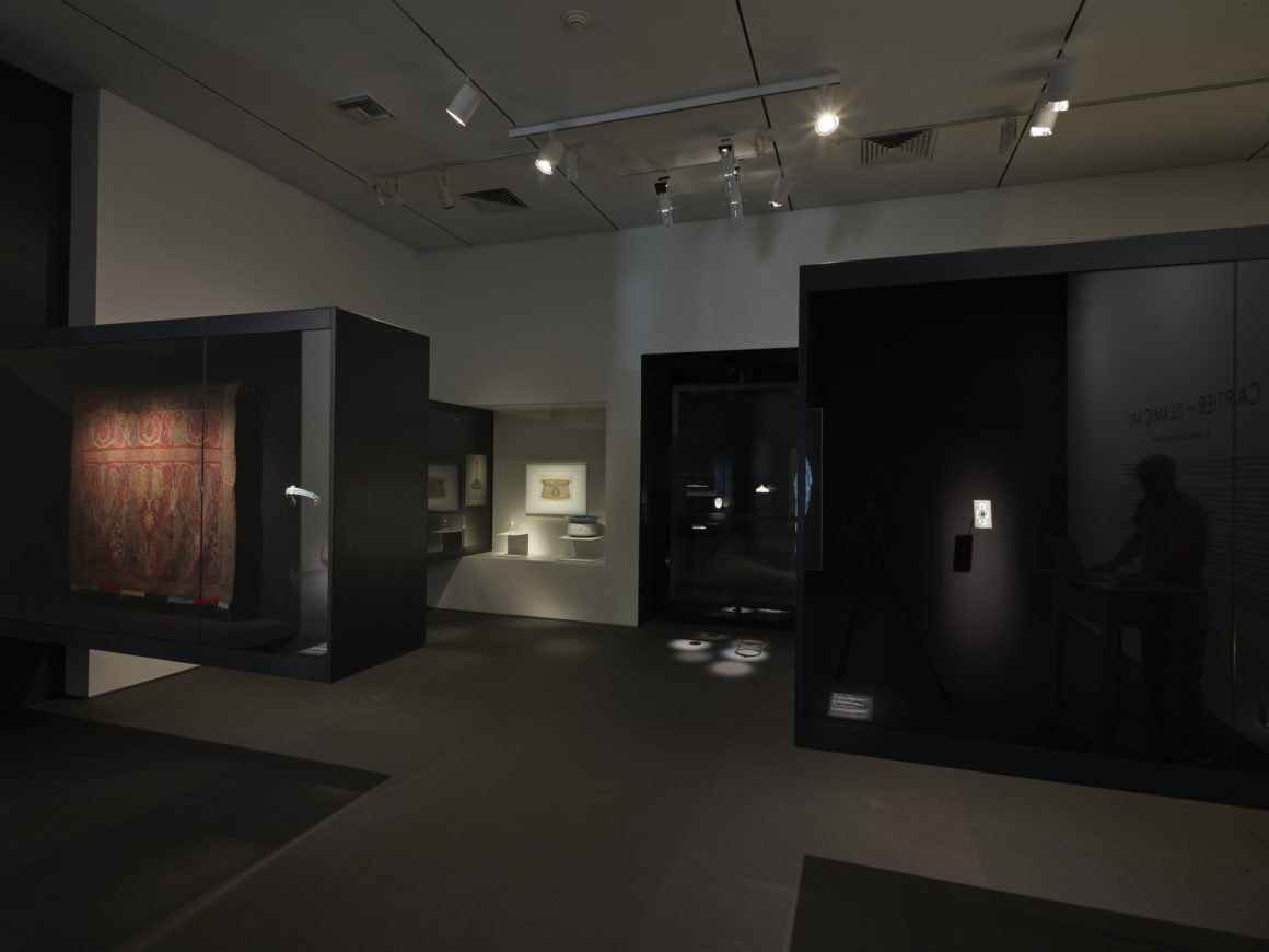 Dark museum exhibit with illuminated display cases showcasing artifacts including pottery and a tapestry.
