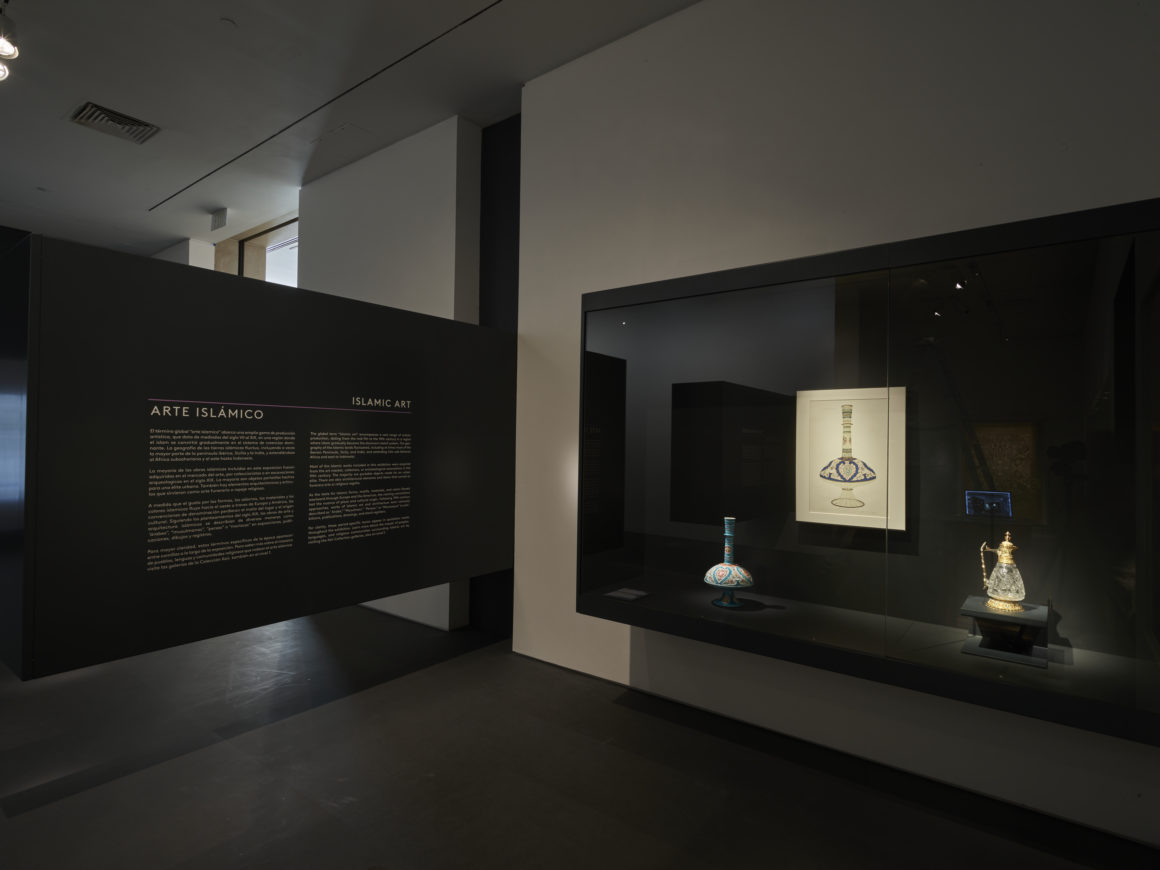 Dimly lit museum exhibit of Islamic art with descriptive text, a display case featuring ceramics, and an illuminated manuscript.