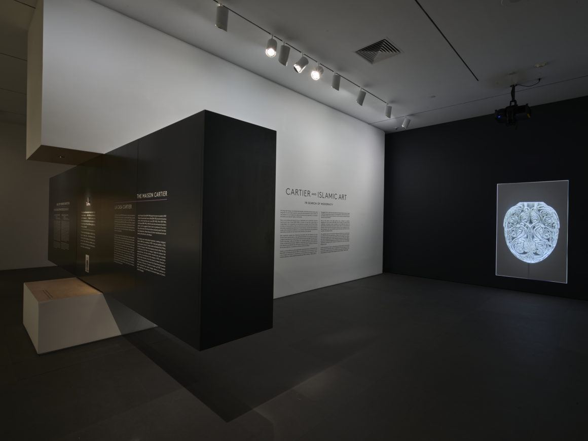 Exhibit room with dark walls displaying text and a glowing framed artwork on the right. Ceiling lights illuminate the space.
