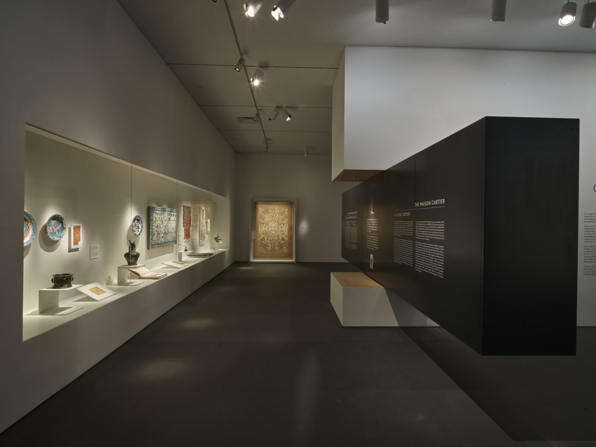 A dimly lit museum exhibit showcases pottery and framed artworks on walls, with informational panels on a black partition.