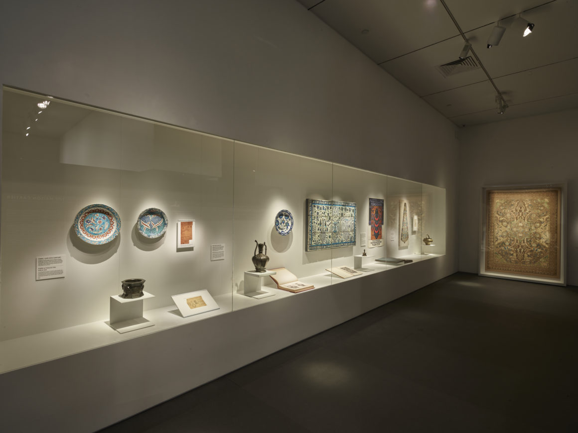 Museum exhibit with illuminated display of ceramics, textiles, and art pieces in a dimly lit room.