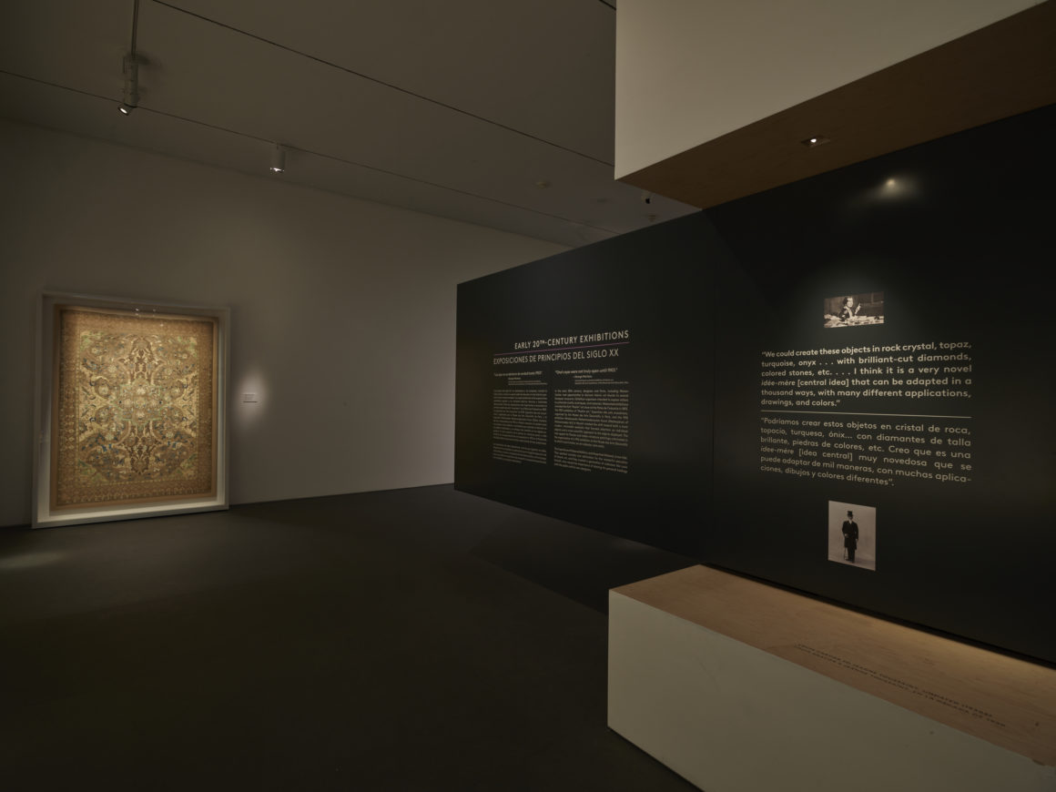 Dimly lit museum exhibit featuring a large tapestry on the left wall and informational text on a dark wall to the right.