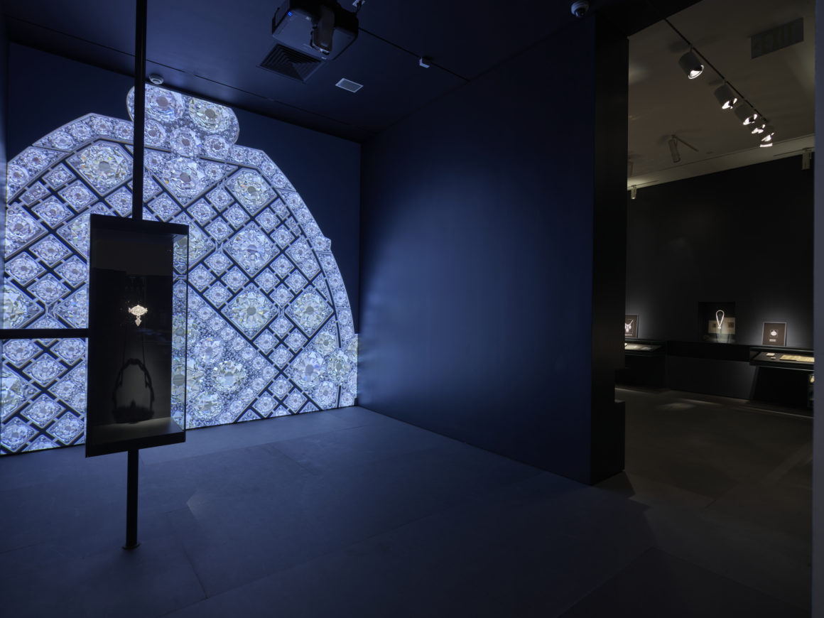 Dimly lit exhibition space with illuminated jewelry display on the left and various cases showcasing jewels on the right.