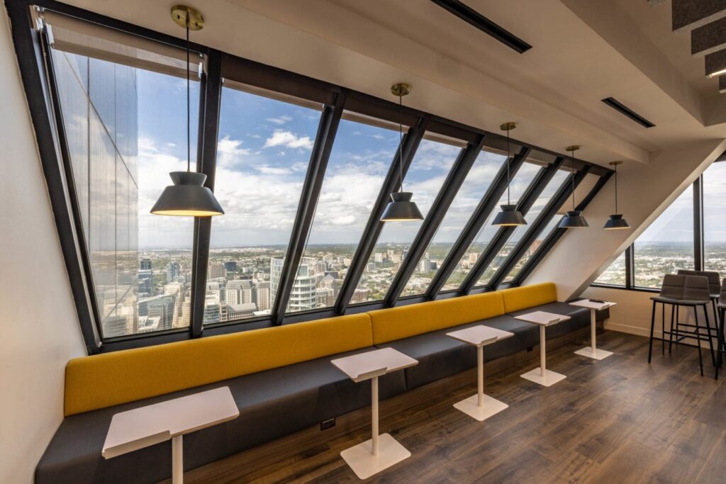 Office lounge with slanted windows offering a city view, yellow bench seating, white tables, and black pendant lights.