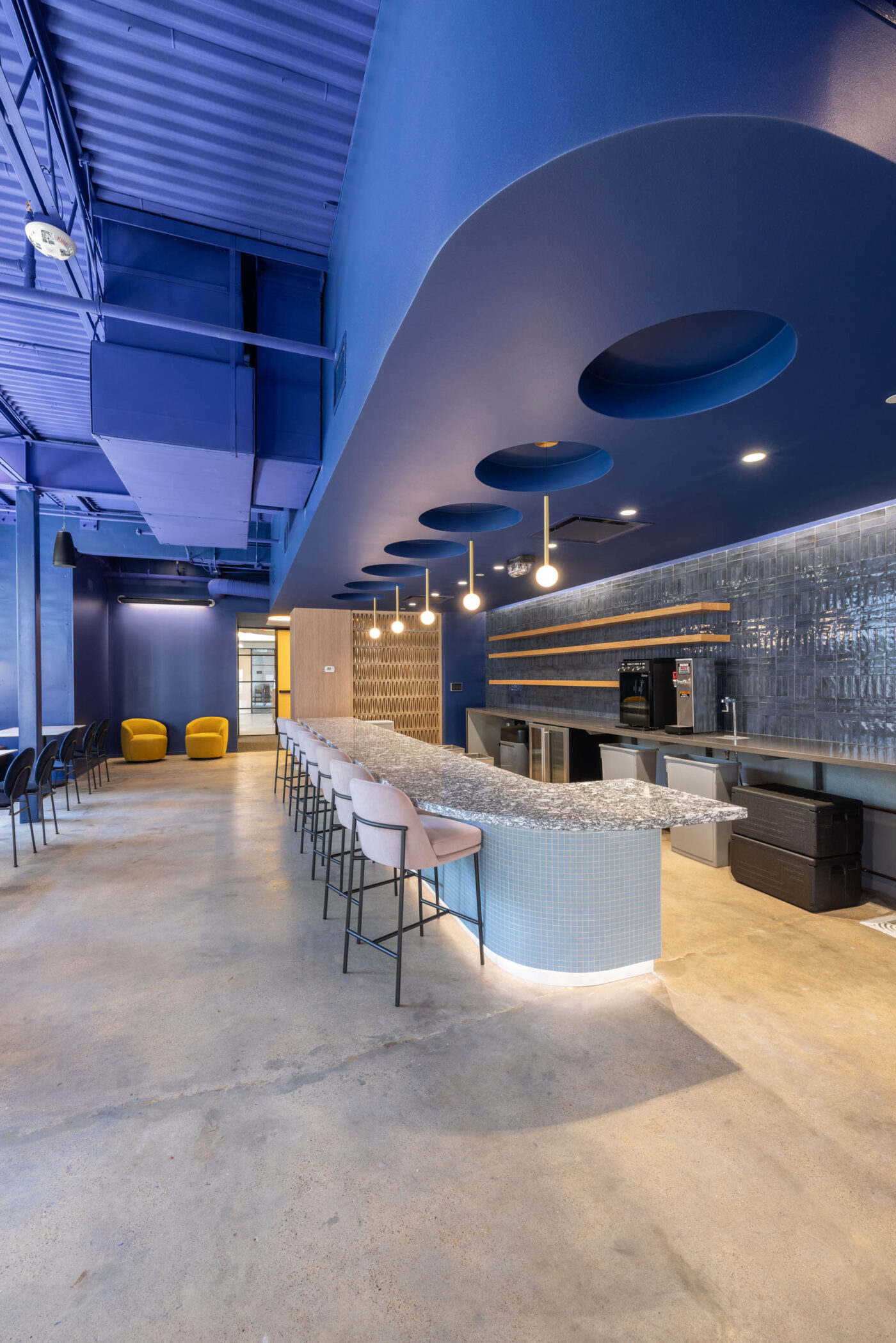 Modern lounge area with a long bar, high stools, yellow chairs, blue walls, and stylish lights for a social hub.