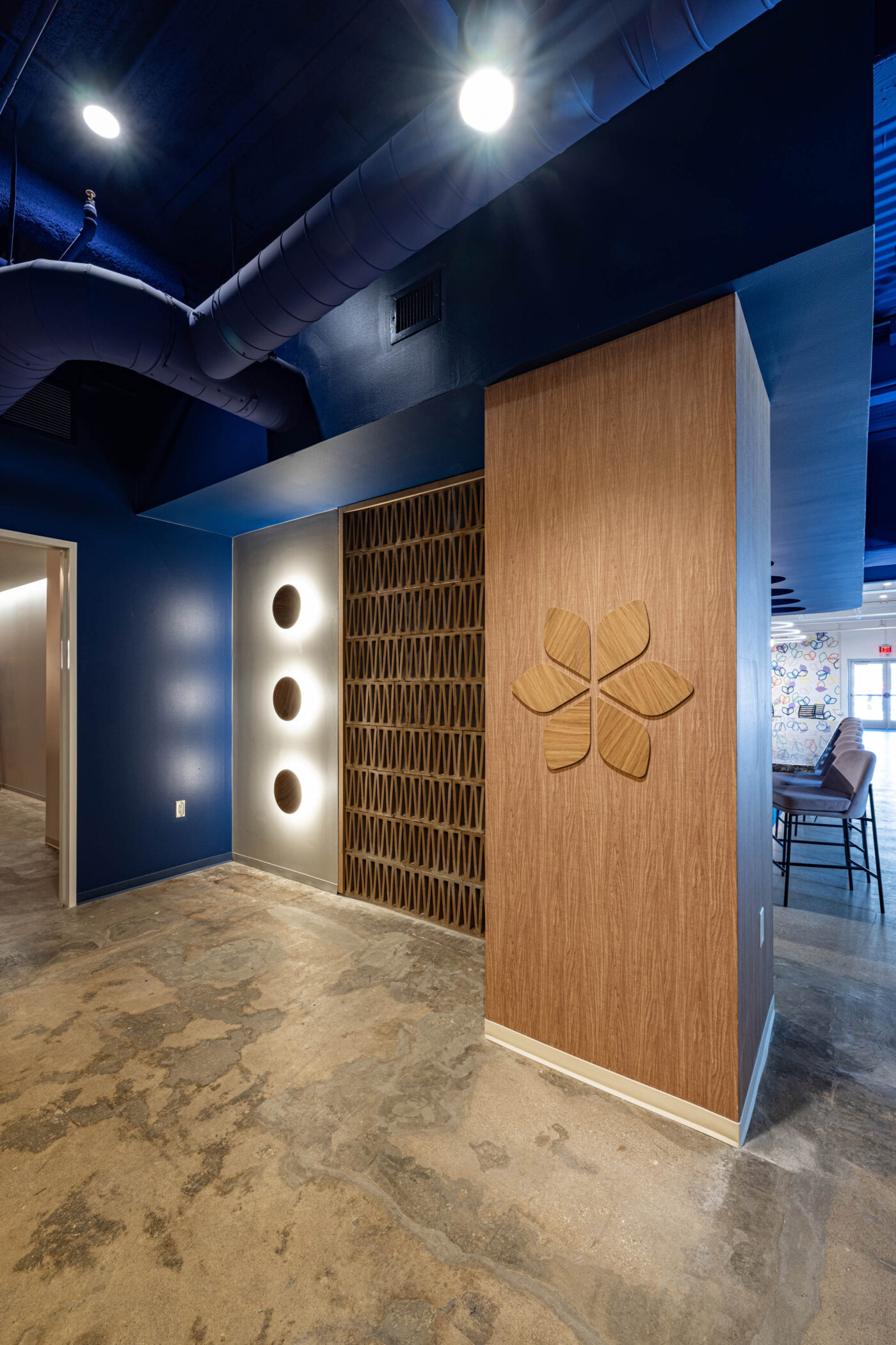 Modern lobby with wooden wall decor, social seating, circular lights on blue wall, exposed ceiling pipes, and concrete flooring.
