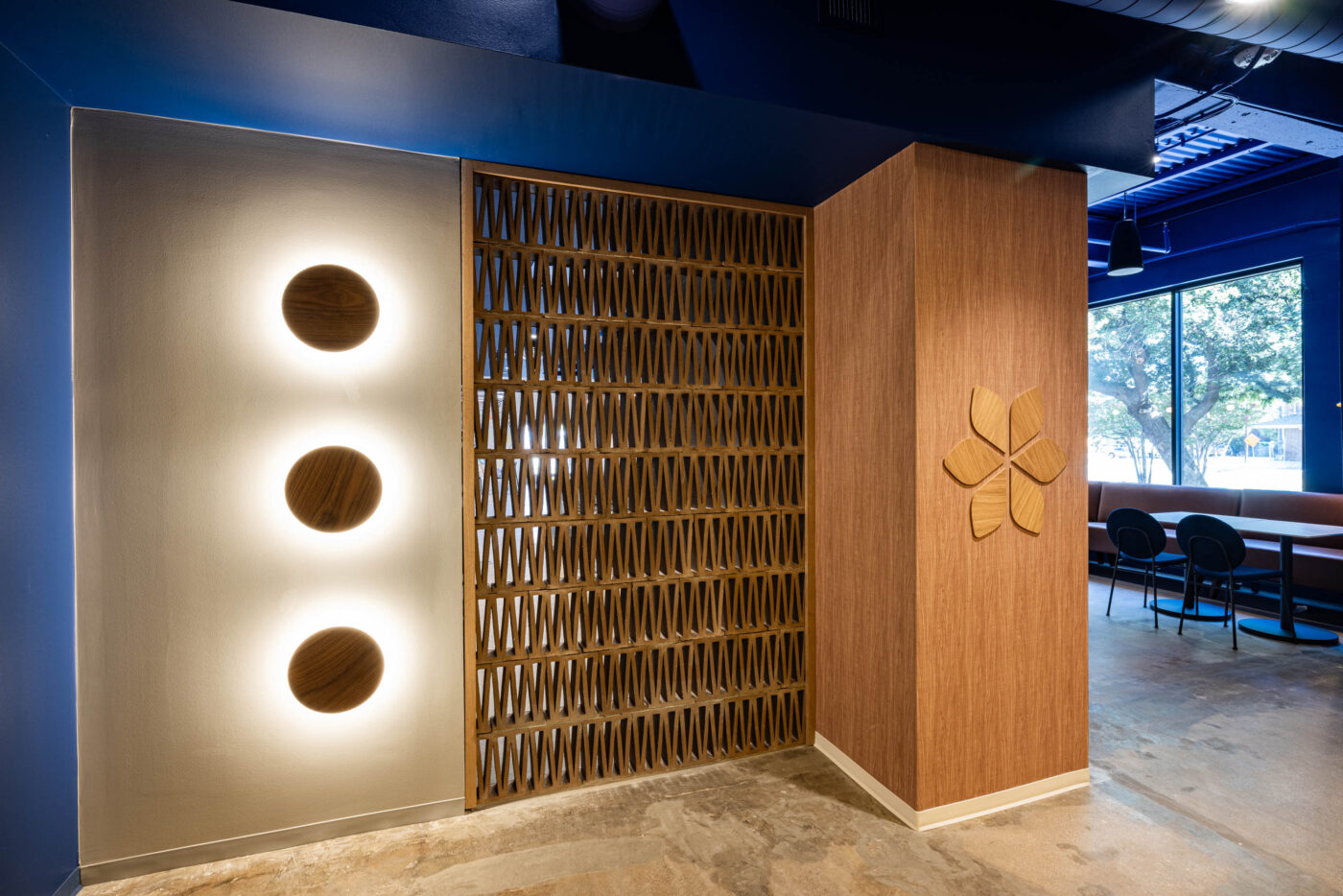 Modern interior with circular wall lights, patterned wooden panel, and floral decor. Large window and tables for a social vibe.