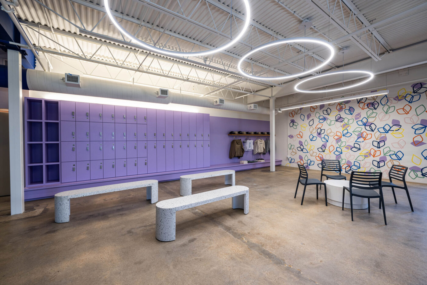 Modern room with purple lockers, benches, and a round table for socializing. Colorful geometric designs and circular lights add vibrancy.