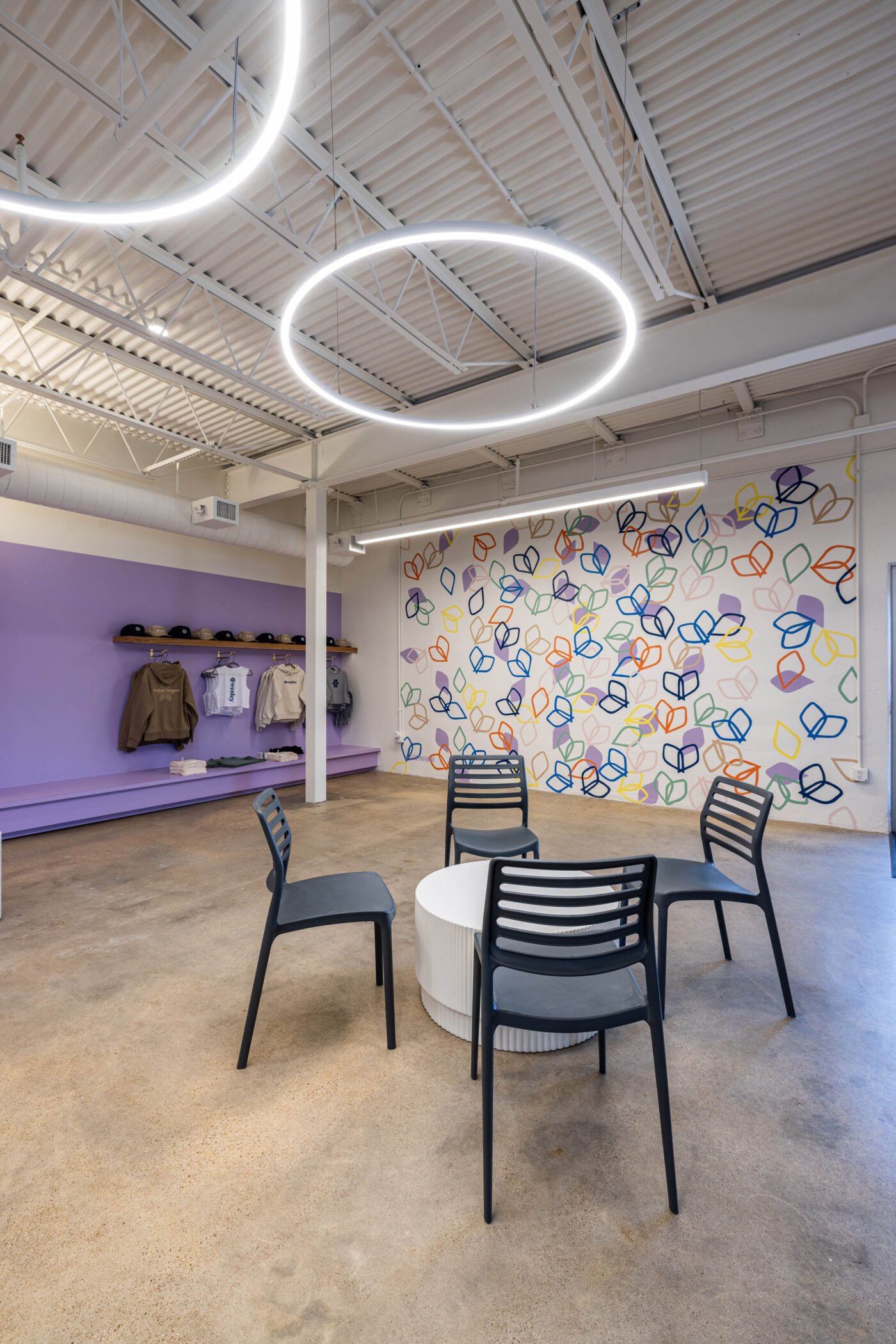 Modern room with circular ceiling lights, a colorful mural, black chairs around a social table, and clothing on shelves.