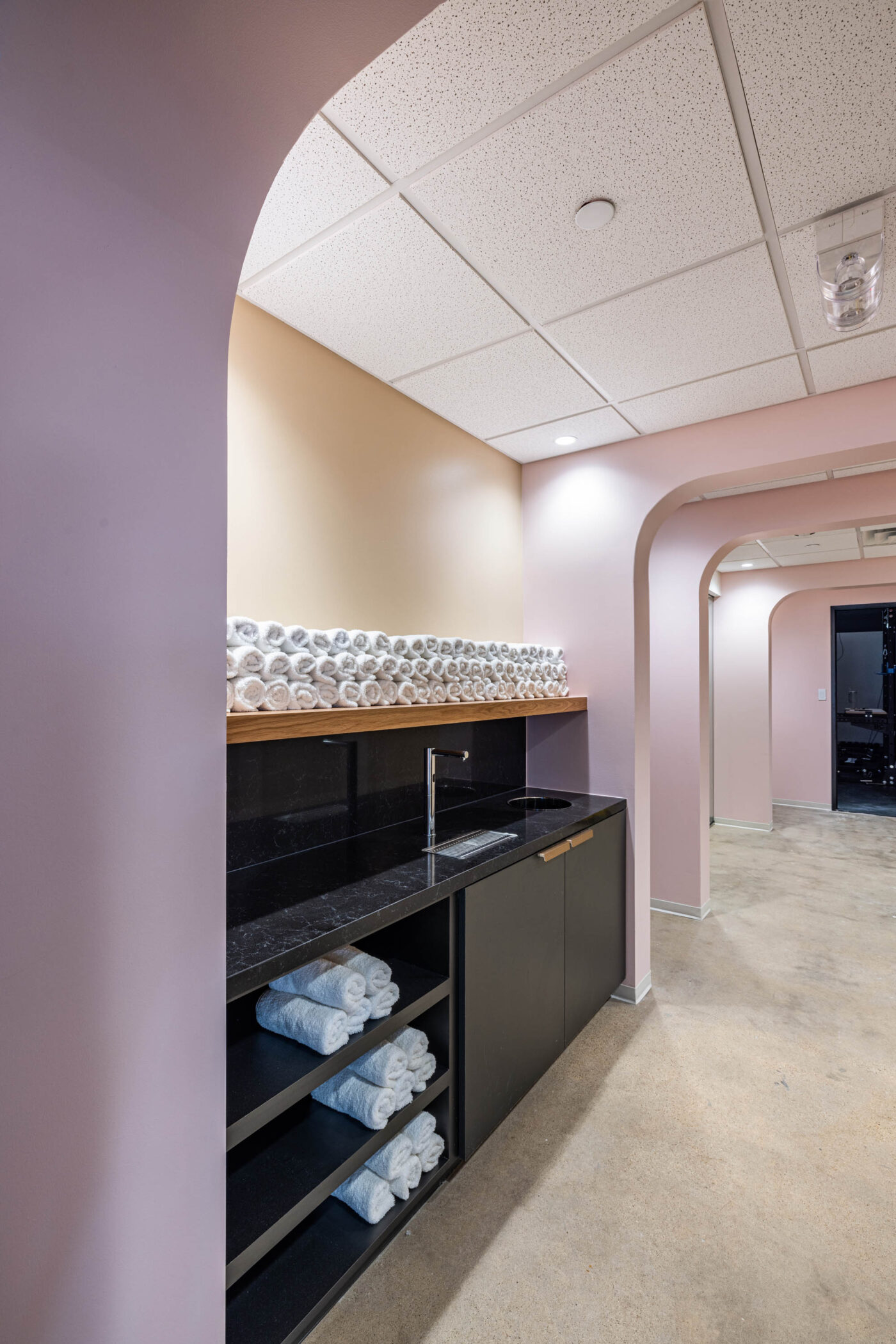 Modern hallway with arched doorways, black counter, sink, and stacked rolled towels on shelves for a sleek fitness vibe.