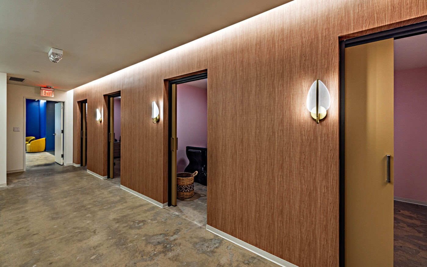 Hallway with wooden walls, four open doors, modern lights, visible pink and blue rooms, concrete floor—perfect for social gatherings.