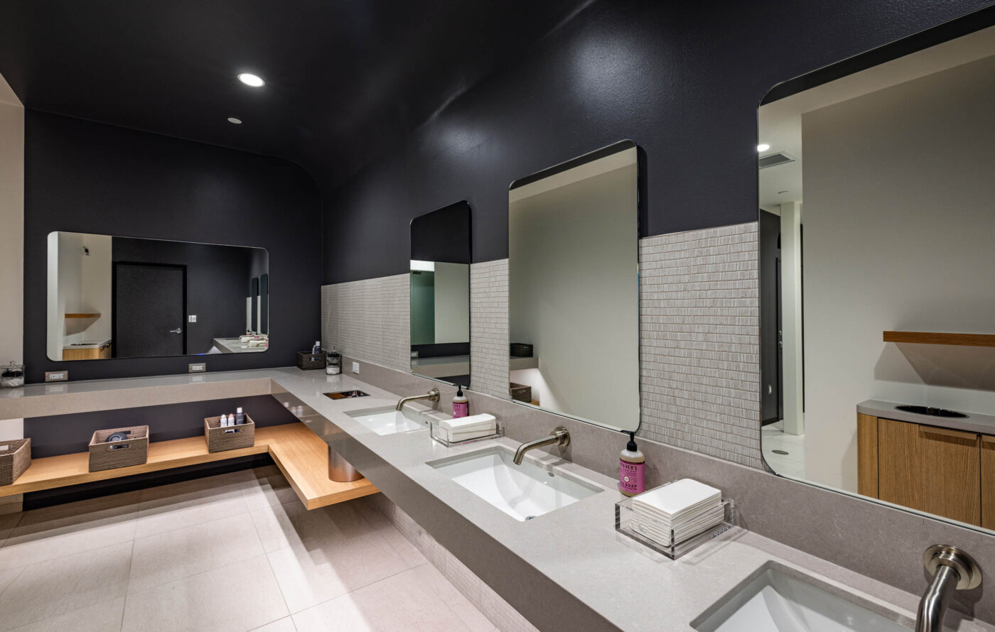 Modern restroom in a fitness center with three sinks, rectangular mirrors, and wooden shelves holding baskets and paper towels.