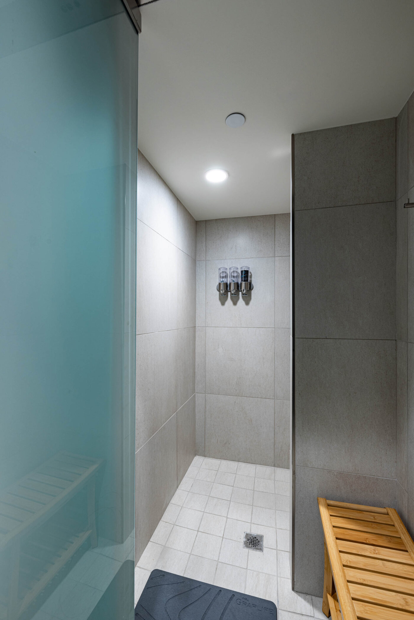 Modern shower area with gray tiles, towel hooks, and a wooden bench—perfect for refreshing post-fitness or social routines.