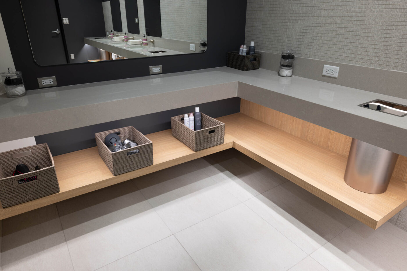 Modern bathroom with corner vanity, woven baskets, and toiletries on a wooden shelf. Large mirror and social space visible.