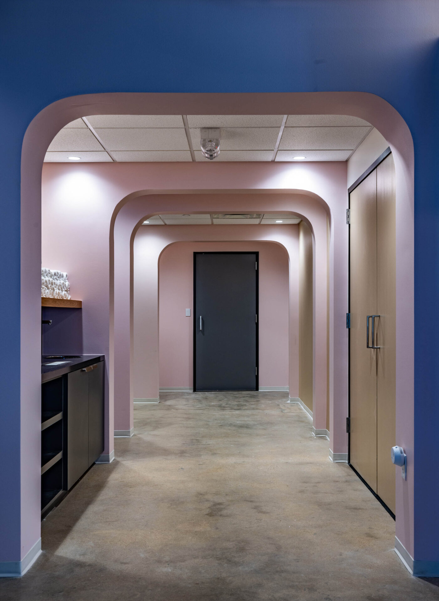 A social hallway with pink walls, arched doorways, a dark door at the end, shelves on the left, and cabinets on the right.