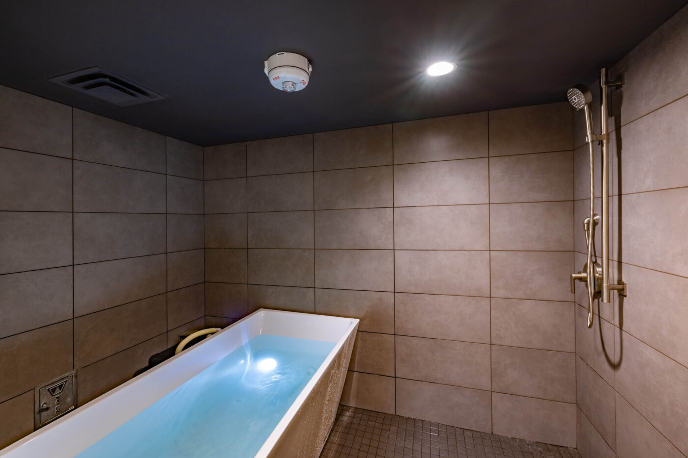 A tiled bathroom boasts a filled bathtub under a ceiling light, ideal for post-fitness relaxation, with a mounted showerhead.