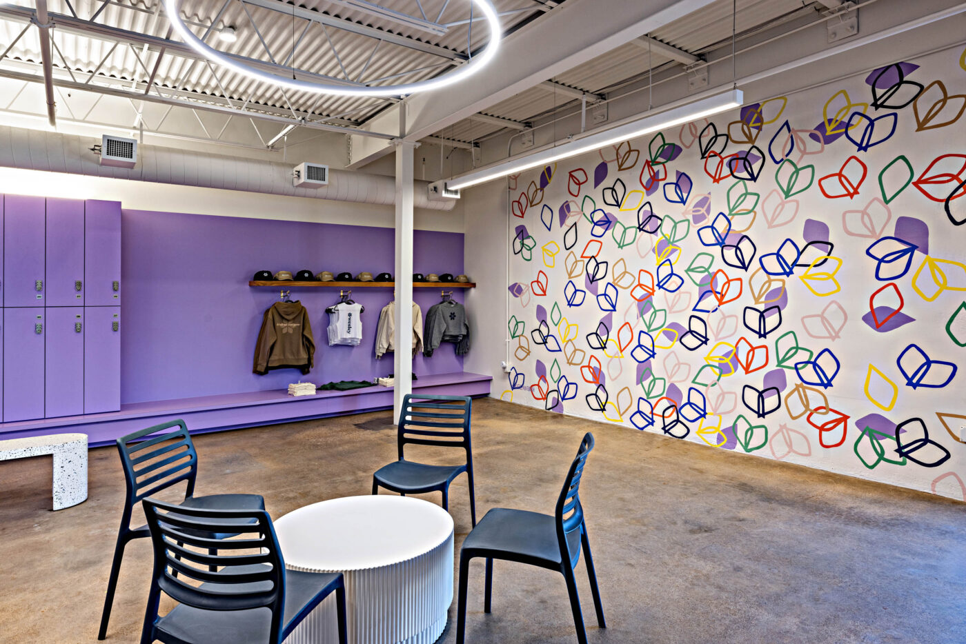 Room with purple lockers, a bench, coats, hats, a colorful leaf-patterned wall, social seating and a round table under a ring light.
