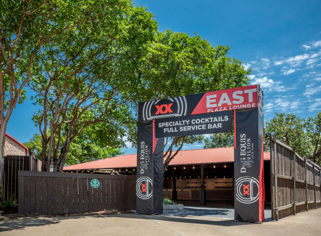 Entrance to East Plaza Lounge with a banner advertising specialty cocktails and full-service bar, surrounded by trees.