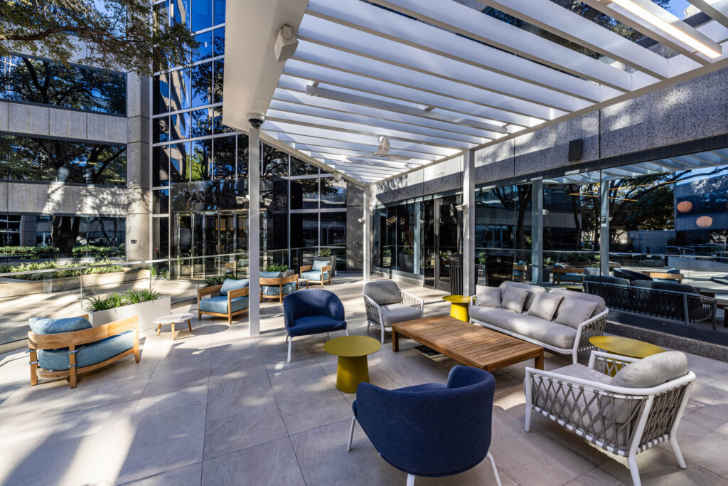 Modern outdoor patio area at Coffee Shop with blue and white chairs, wooden tables, and glass reflections under a slatted roof.