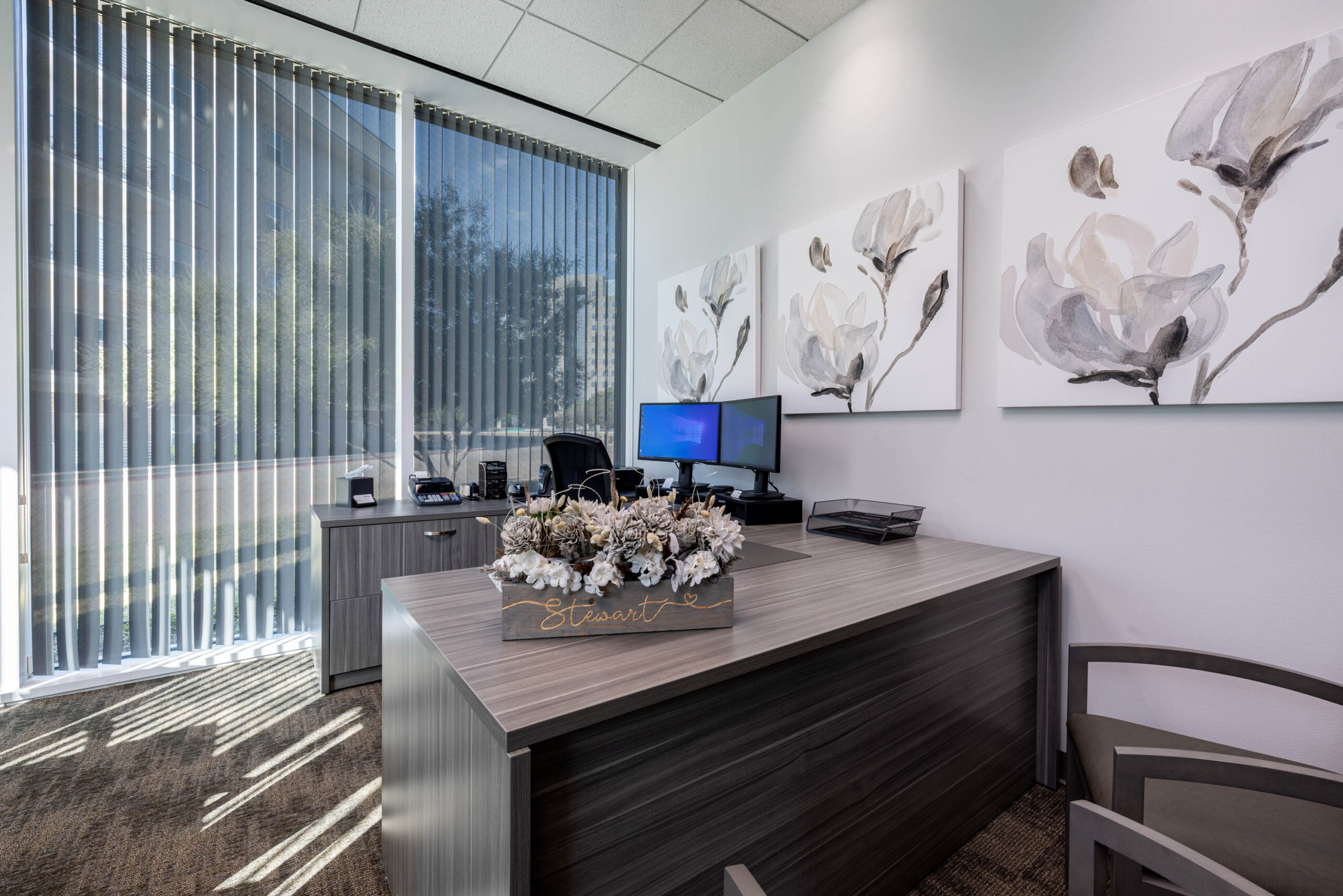 Modern office with a wooden desk, chairs, Stewart Title artwork, and vertical blinds showing a sunny tollway view outside.