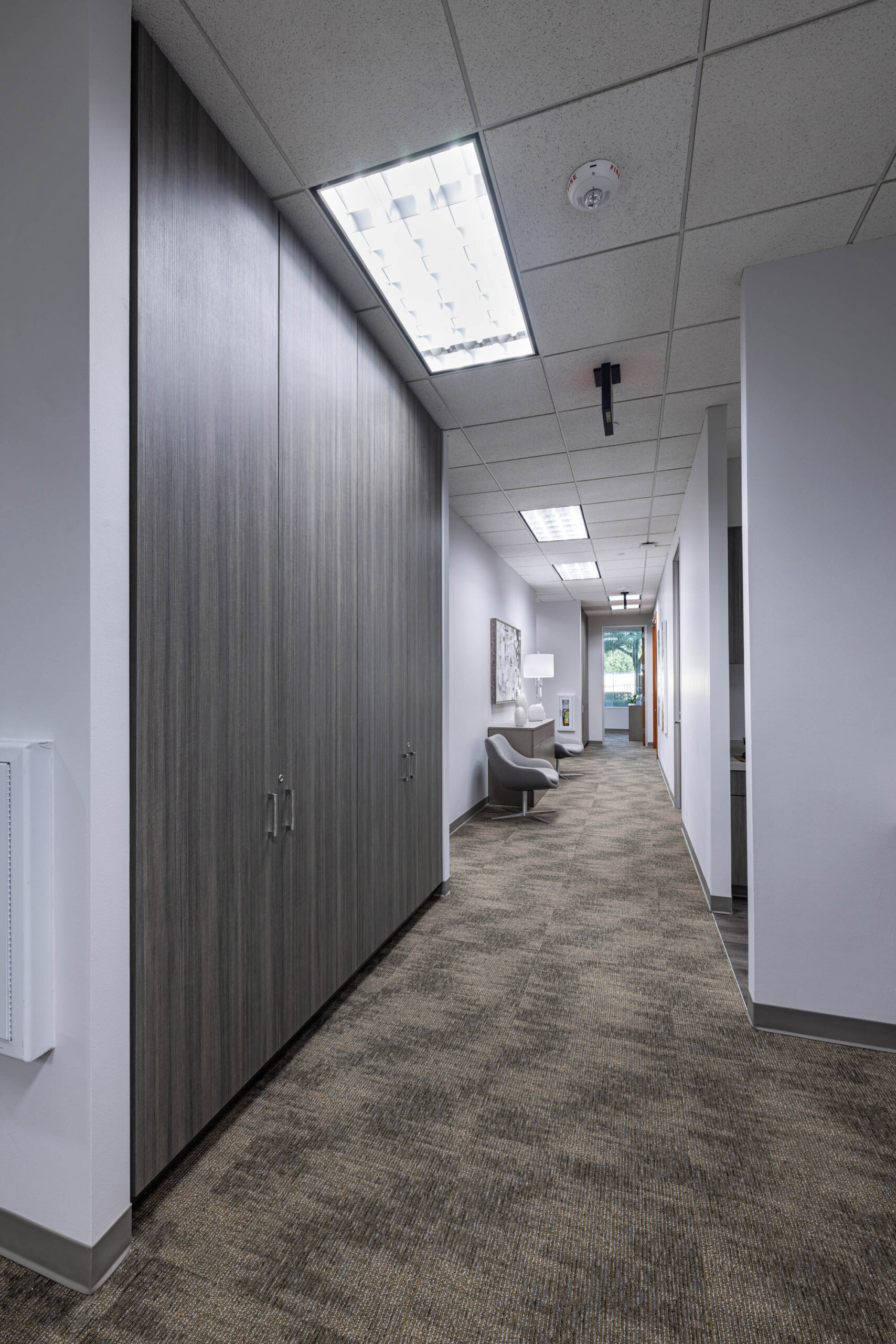 Modern office hallway with wood-panel storage, patterned carpet, and a chair by Stewart Title near artwork and a window.