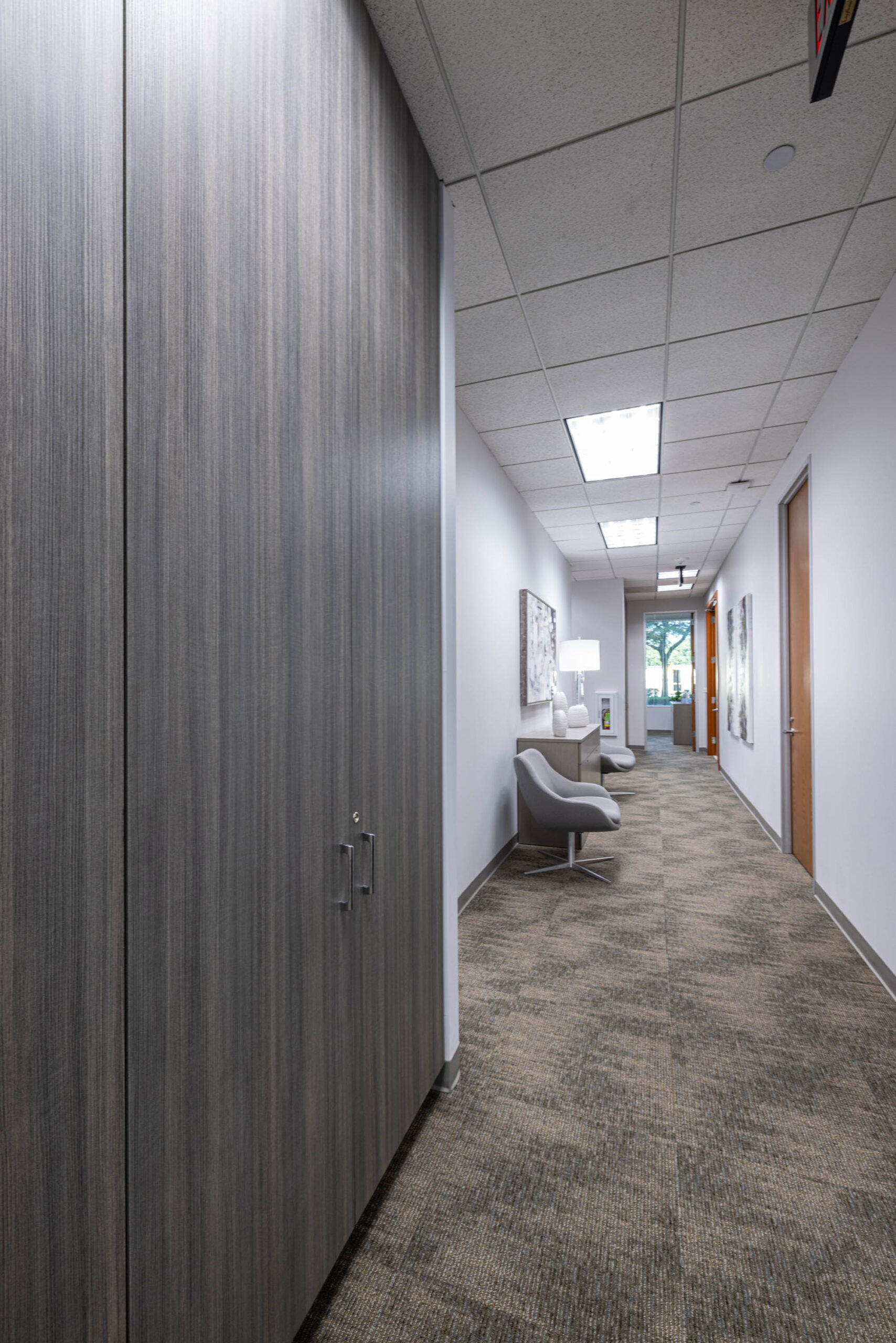 A modern office hallway with gray carpeting, Stewart Title paneling, chairs, artwork on walls, and ceiling lights.