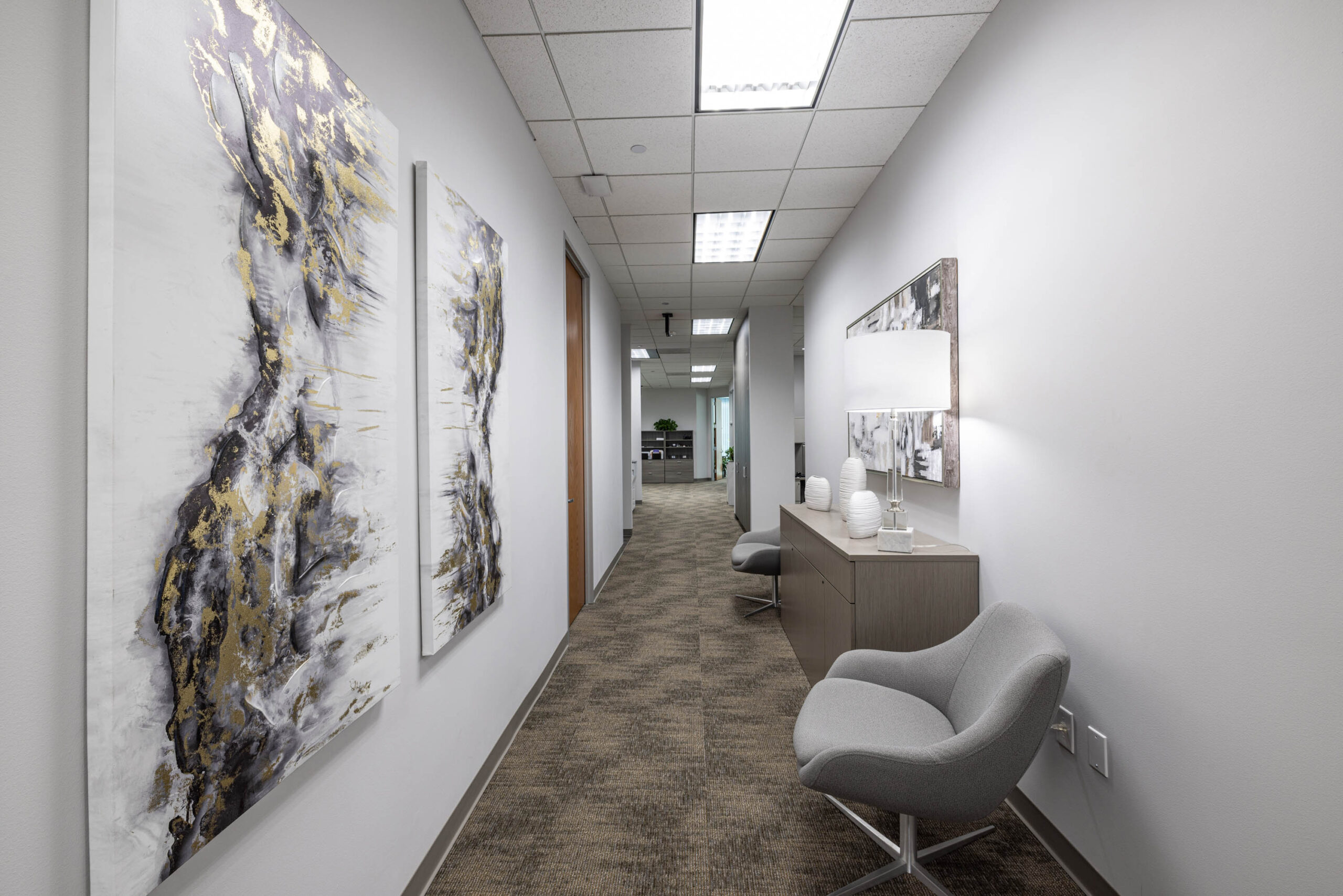 Modern office hallway with abstract wall art, a Stewart Title console table, and a gray chair on a carpeted floor.