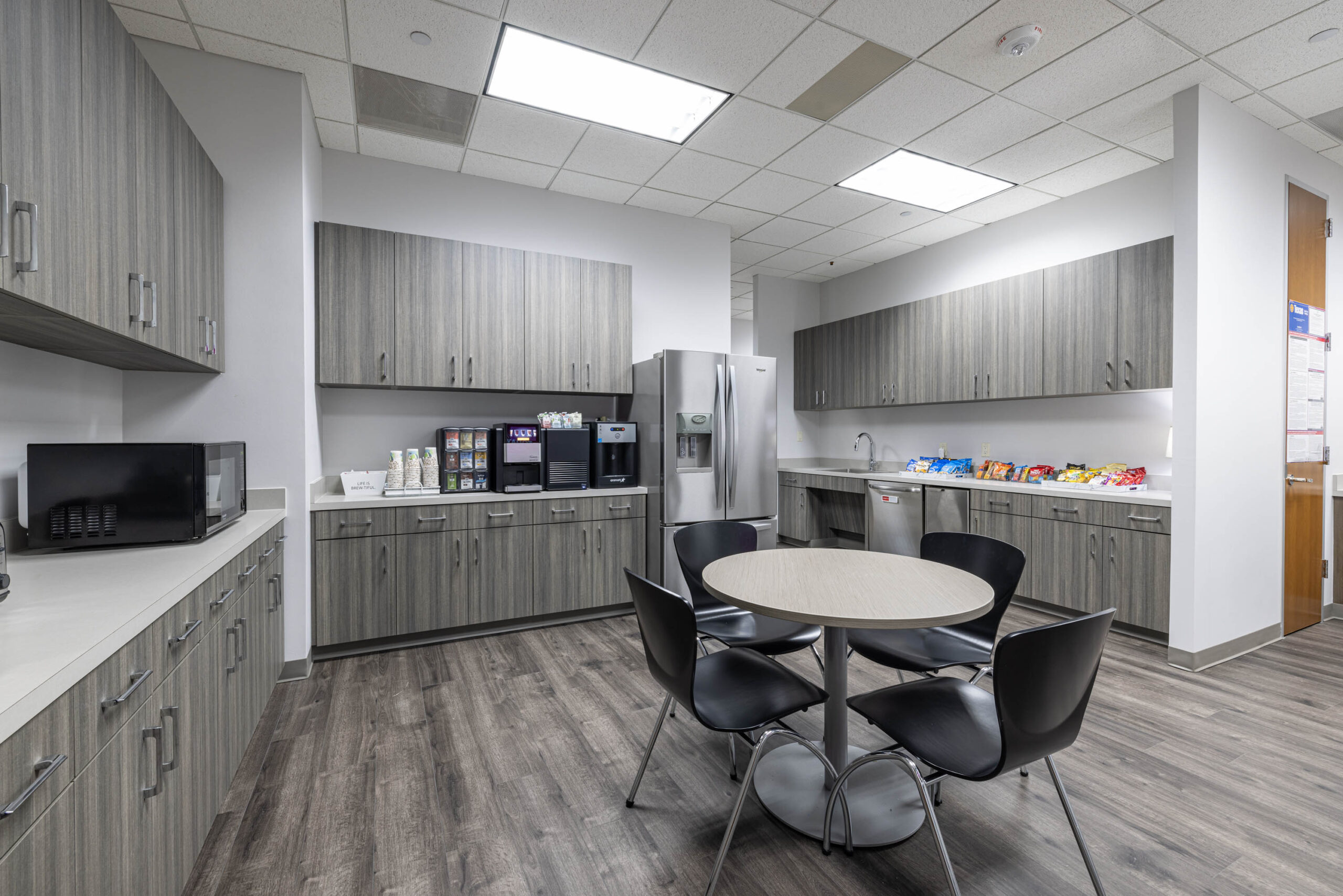 Modern office kitchen near Tollway with gray cabinets, round table, black chairs, stainless fridge, and snack area.