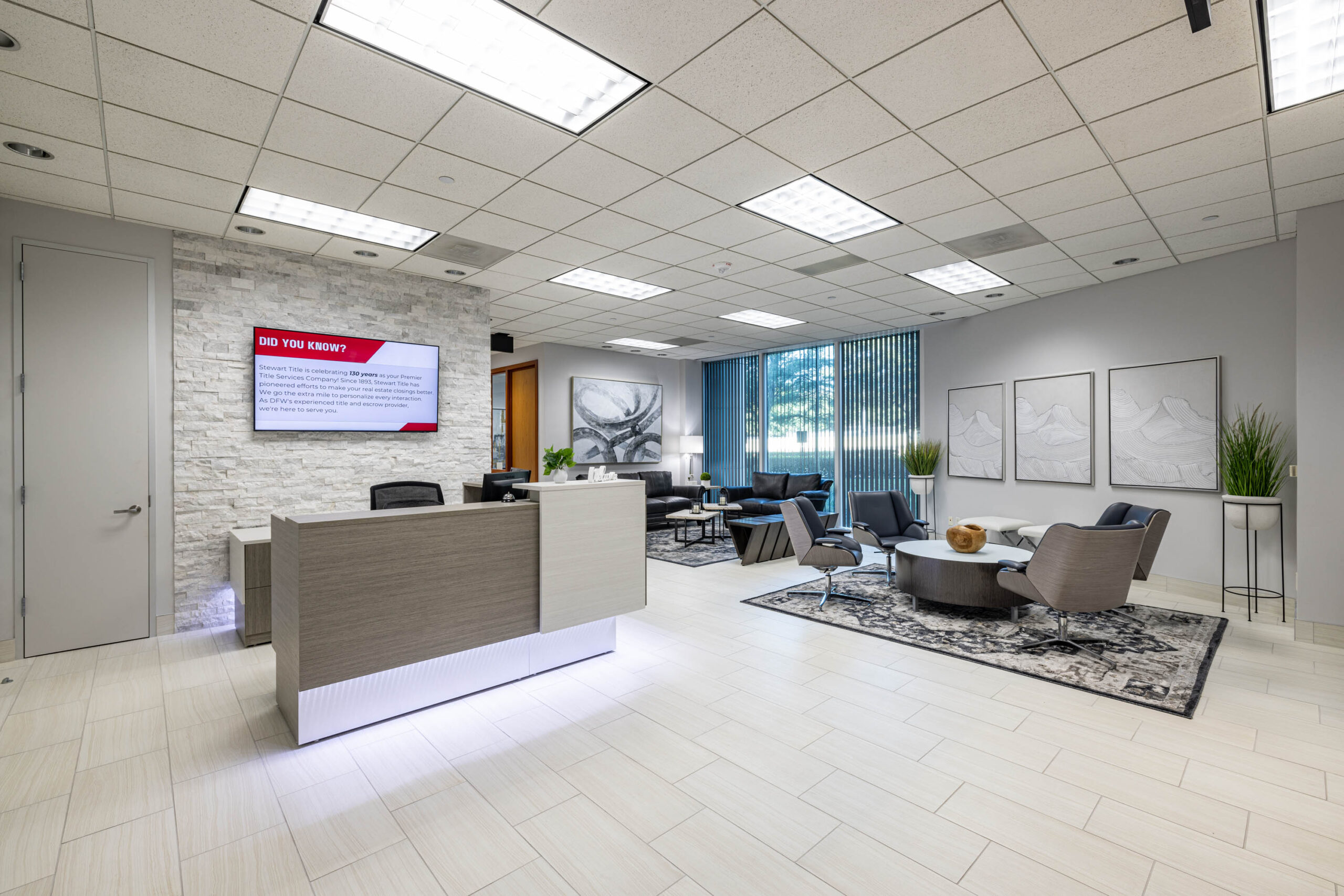 Modern office reception area with a central desk, seating, and decorative plants. Tollway access and bright ceiling panel lighting.