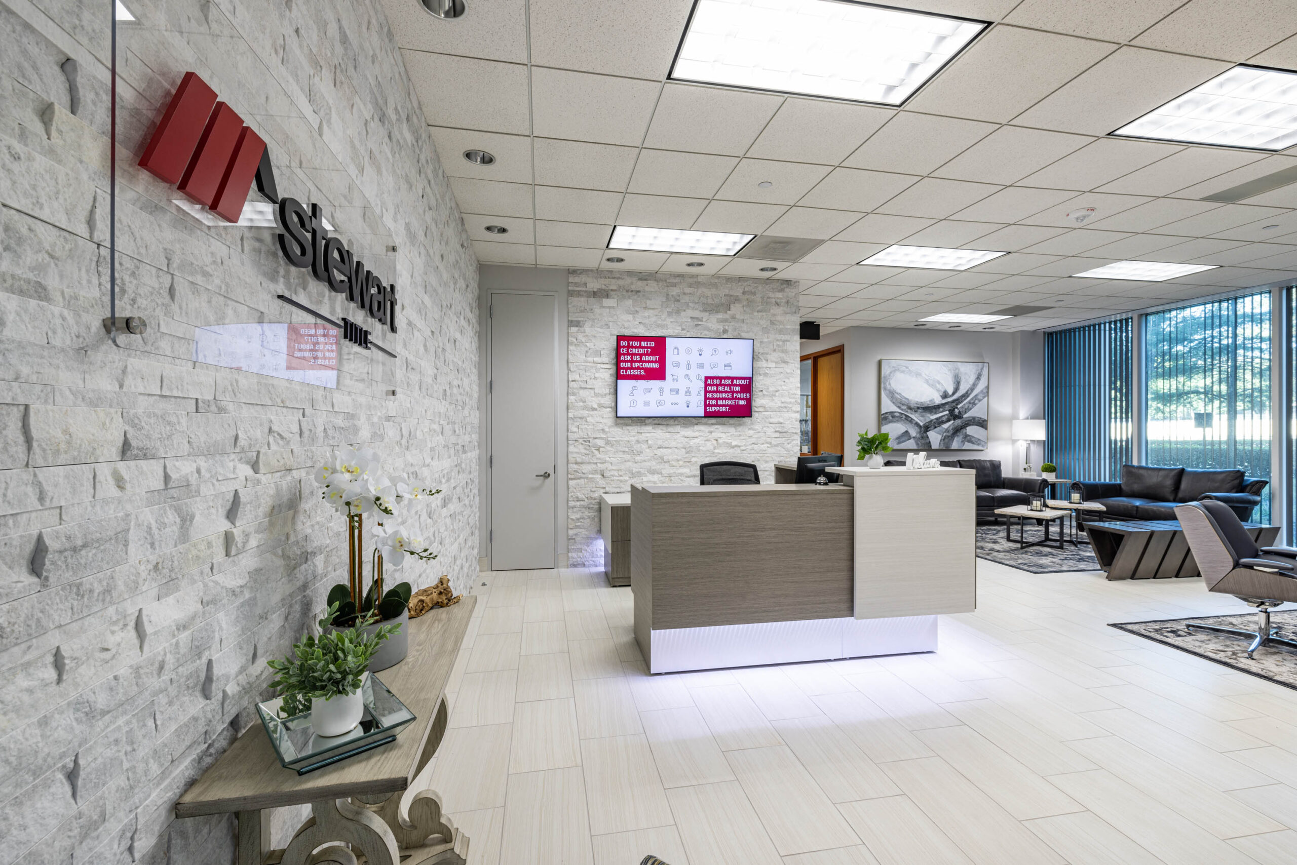Modern office lobby with a reception desk, Stewart Title signage, seating area, potted plants, and large windows with blinds.