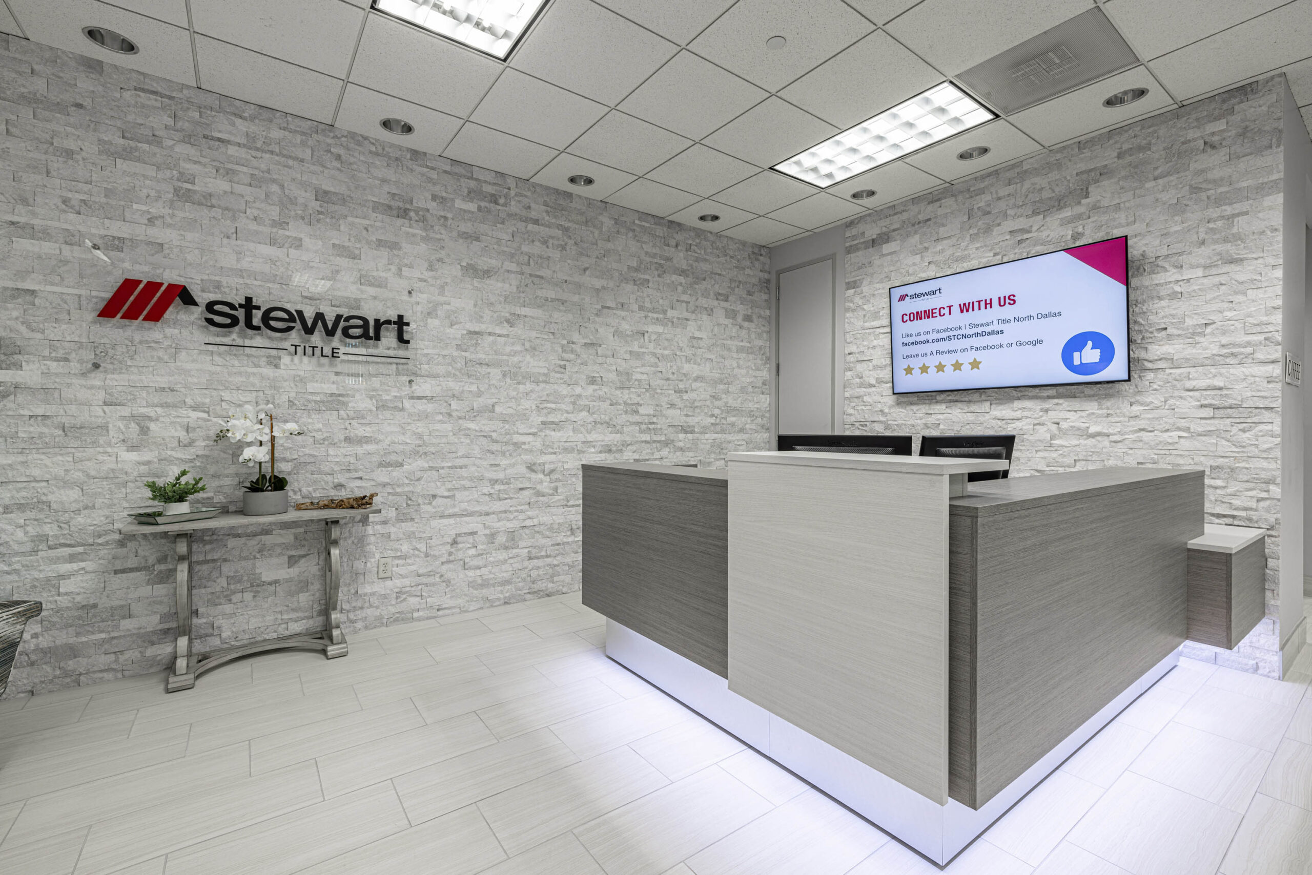 A modern reception area with a gray desk, wall-mounted monitor, and Stewart Title logo on a light brick Tollway wall.
