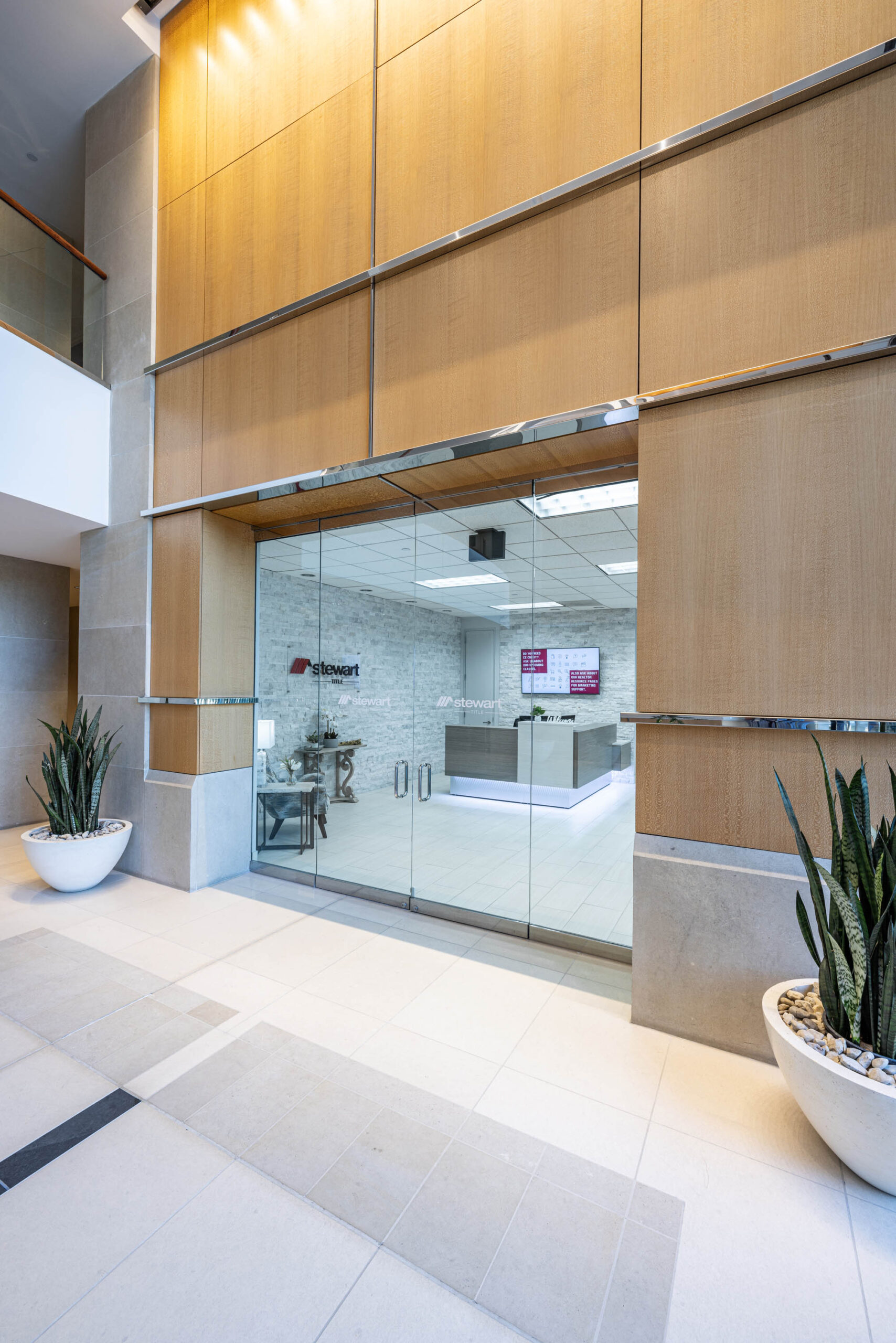 Modern lobby entrance with glass double doors, Tollway views, large potted plants, light wood panels, and a bright reception area.